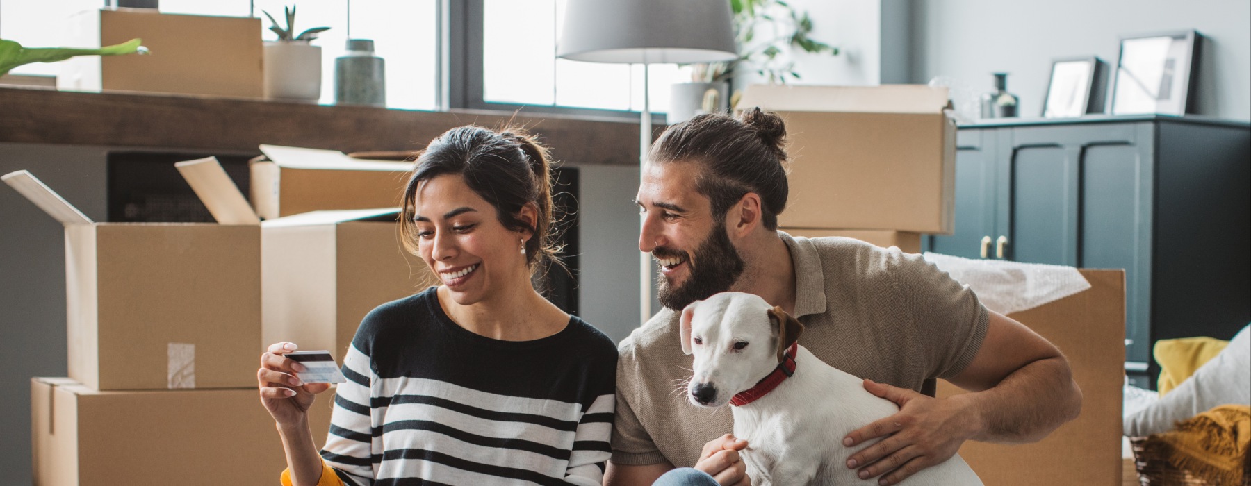A smiling couple sitting on the floor of their new pet-friendly apartment surrounded by moving boxes, using a laptop and credit card with their dog.